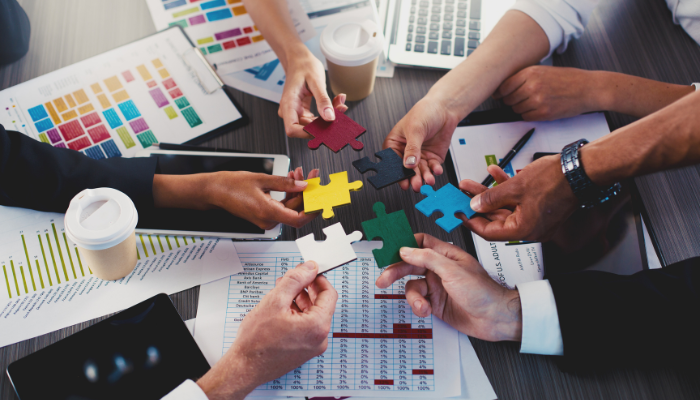 Hands holding multicoloured jigsaw puzzle pieces over a desk cluttered with business papers.