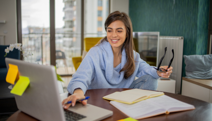 A businesswoman sitting in front of some books holding a pair of glasses while keying on a laptop while holding a pen in her hand.