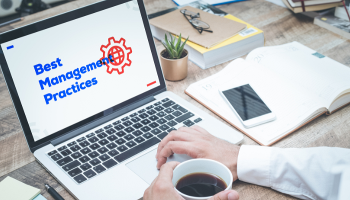 A person holding a cup of coffee working on a laptop wit the words "Best Management Practices" on the screen.